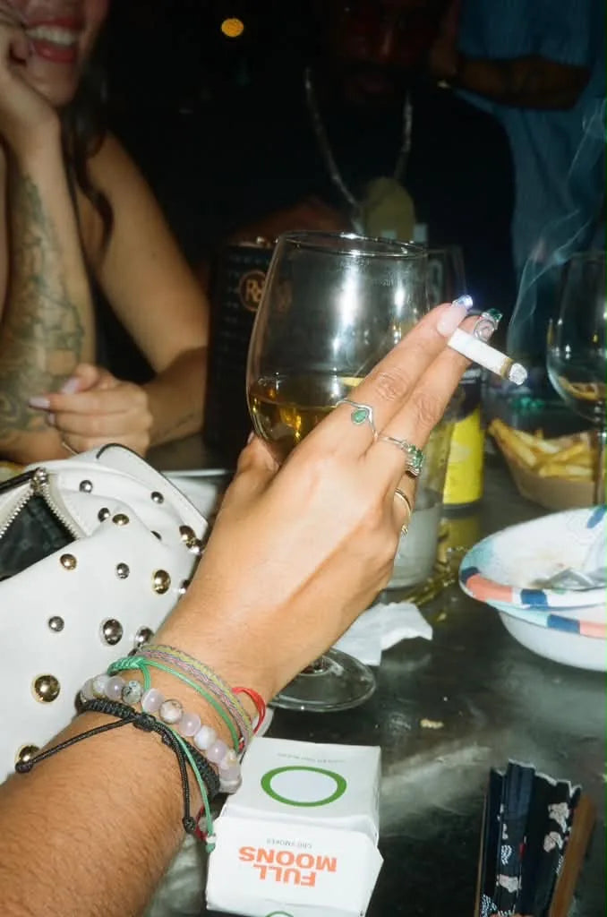 A hand holding a hemp cigarette and a glass of wine at a night table, a Full Moons pack below